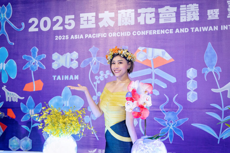 主持人 Yuki 身穿黃色禮服，以蘭花女神造型，在 2025 亞太蘭花會議的紫色背板前，優雅地與蘭花合影。