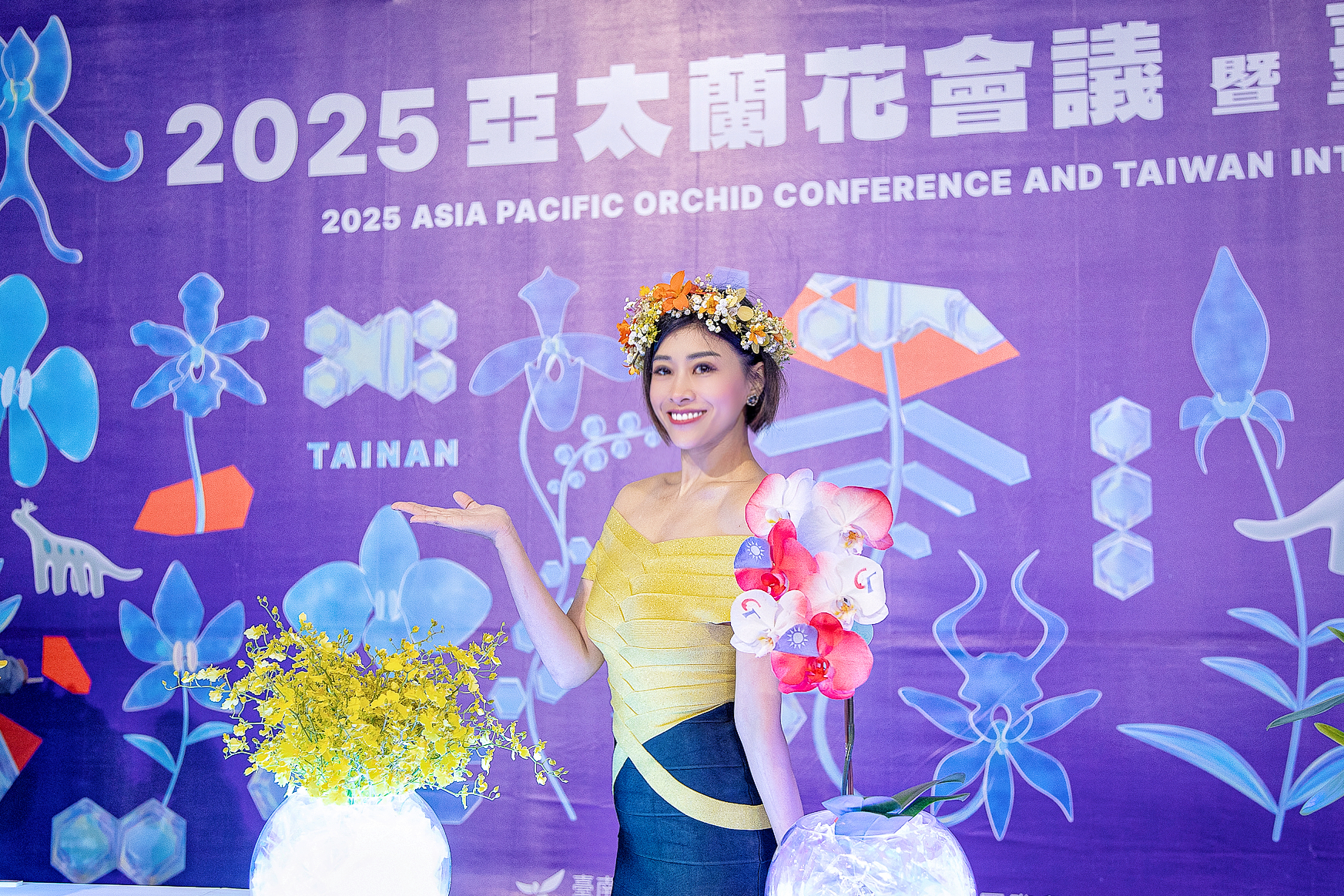 主持人 Yuki 身穿黃色禮服,以蘭花女神造型,在 2025 亞太蘭花會議的紫色背板前,優雅地與蘭花合影。