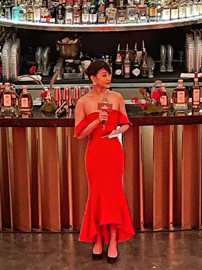 專業雙語主持人 Yuki 身穿紅色魚尾長禮服,在 HINE 干邑品牌活動的吧台前,手持麥克風專注地進行主持工作。